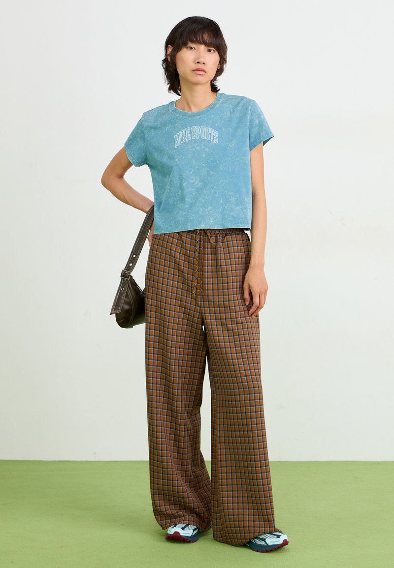Lichtblauw cropped t-shirt met "Nike Sports" print, gecombineerd met bruine geruite wijde broeken en sneakers met rode accenten.