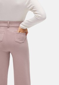 Pantalones de tiro alto rosa con una textura suave, que cuentan con dos bolsillos traseros. La modelo lleva una camiseta blanca de manga larga con canalé.