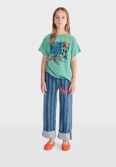 Fille aux longs cheveux portant un t-shirt graphique vert, un jean rayé retroussé avec texte rouge, et des chaussures orange sans lacets, debout devant un fond uni.