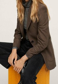 Blazer marrón de espiga con solapas de pico, cierre de un solo botón y tejido texturizado, combinado con un suéter de cuello alto gris y pantalones negros.