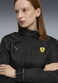 Jachetă neagră cu guler înalt, linii gri și logo-ul Ferrari pe piept. Materialul pare texturat și structurat, cu un detaliu la manșetă cu nasturi.
