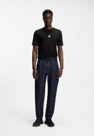 Man staat en draagt een zwart T-shirt met opdruk, donkerblauwe spijkerbroek met riem en zwarte sneakers, tegen een effen witte achtergrond.