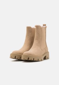 Botine bej din suede, cu o căptușeală moale, panouri laterale elastice și o talpă robustă, cu striații. Dispune de o tabără la spate pentru a fi ușor de purtat.