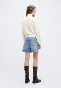 Γυναίκα που φοράει λευκό πουλόβερ με ψηλό γιακά, ανοιχτό μπλε τζιν σορτς και μαύρες μπότες μέχρι τη μέση της γάμπας, σε πλευρική όψη με λευκό φόντο.