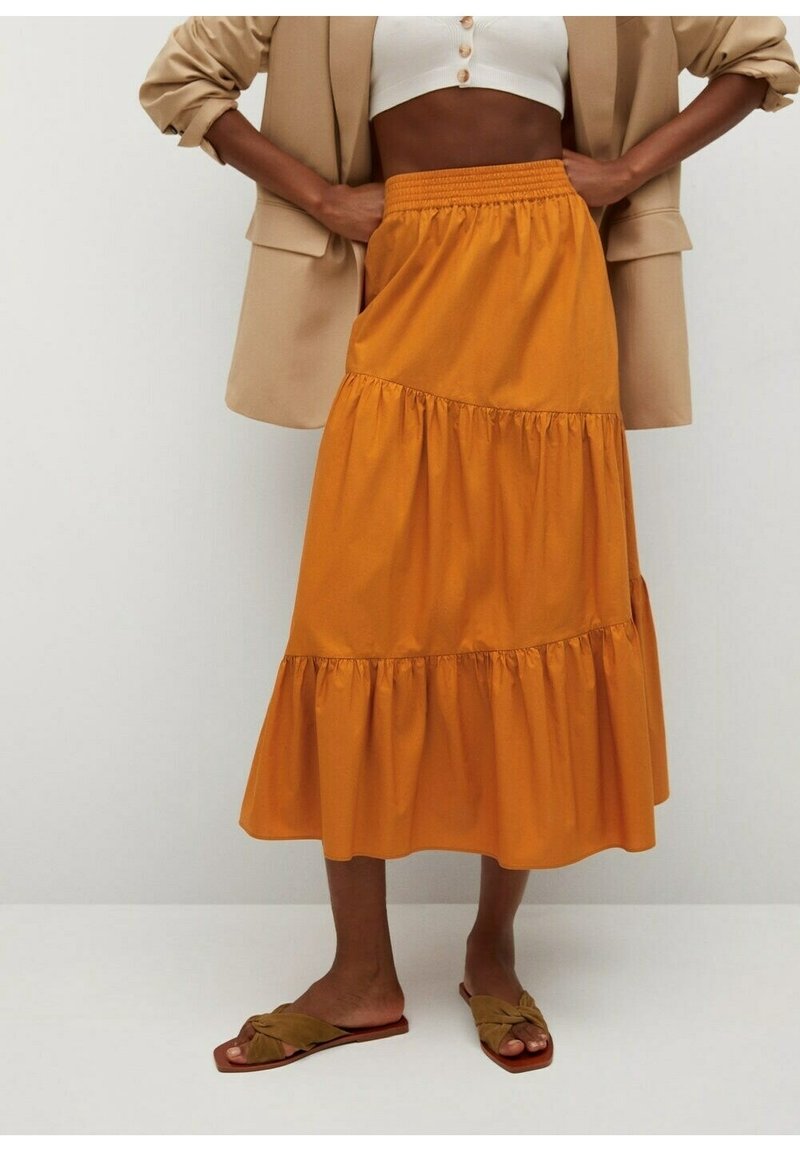 Femme portant une jupe midi à volants jaune moutarde avec une taille élastique, un top blanc court, un blazer beige et des sandales plates couleur tan.