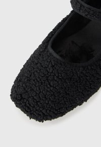 Bailarinas negras hechas de un material texturizado y esponjoso, con una punta redonda y una correa elástica que cruza la apertura del pie.