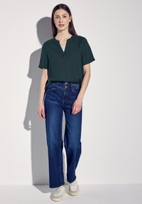 Top corto a maniche corte di colore verde scuro con scollatura a V, abbinato a jeans in denim blu larghi e sneaker bianche. Tessuto morbido e vestibilità rilassata.