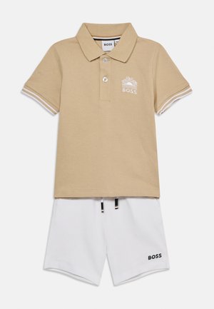Beige poloshirt med hvit logo og knapper, kombinert med hvite shorts med svarte snøringer og en svart logo på benet.