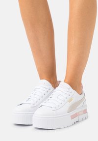 Vita plattformssneakers med ovan i läder, som har rosa accenter och en guldfärgad logga. Texturerad sula med vertikala linjer för bra grepp.