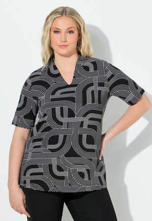 Kvinde med blondt hår iført sort-hvid kortærmet top med geometrisk mønster, stående med den ene hånd på hoften mod en hvid baggrund.