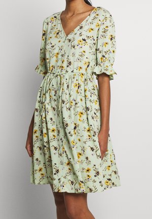 Frau trägt ein hellgrünes Blumenkleid mit kurzen Puffärmeln, V-Ausschnitt, Knöpfen vorne und einem Taillengürtel, steht vor einem schlichten Hintergrund.