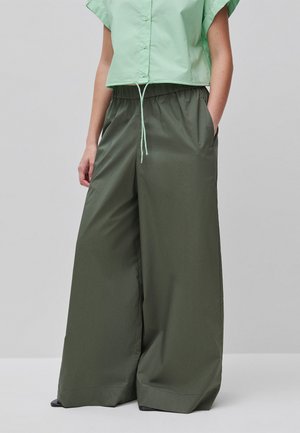Personne portant un pantalon large vert foncé avec taille élastique et une chemise courte boutonnée vert clair, mains dans les poches.