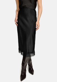 Schwarzer Satinhrock mit Spitzenbesatz am Saum. Glatte Textur und gerade Silhouette; kombiniert mit spitzen, dunkelbraunen Lederstiefeln.
