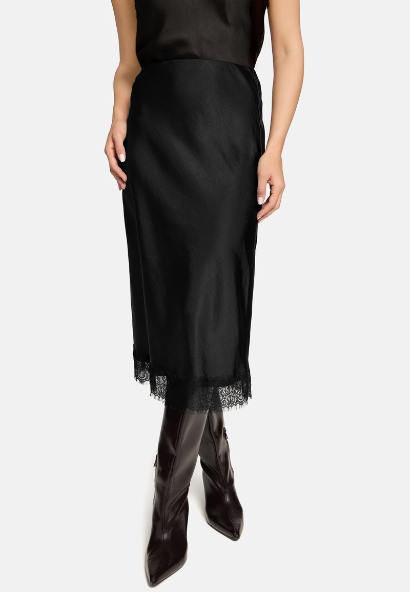 Schwarzer Satinhrock mit Spitzenbesatz am Saum. Glatte Textur und gerade Silhouette; kombiniert mit spitzen, dunkelbraunen Lederstiefeln.