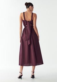 Burgundijska lanena obleka s širokimi nabranoščenimi naramnicami, odprtim hrbtom in detajlom v obliki pentlje pri pasu; do gležnjev dolga s prosto prileganje. Črne sandale s peto.