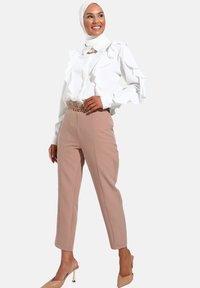 Hvid bluse med flæser og lange ærmer, parret med lyserøde skræddersyede bukser og et guld kædebælte. Beige hæle fuldender looket.