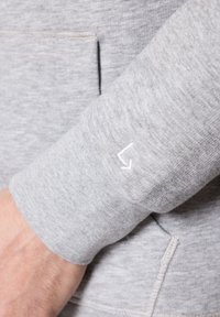 Jasnoszara tkanina bluzy z teksturowanym wyglądem. Rękaw ma mały, biały detal z logo. Zawiera kieszeń z przodu.