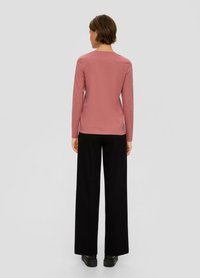 Osoba stojąca tyłem, ubrana w długi rękaw w kolorze przybrudzonego różu, czarne spodnie o szerokich nogawkach i czarne buty na jednolitym szarym tle.