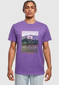 Purpurs t-krekls ar taisnstūra grafiku, kurā attēlots basketbola laukums ar kalniem un palmu kokiem, teksts saka: "sajūti karstumu."