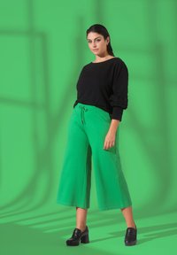 Pantalons larges verts avec une taille élastique à cordon, associés à un pull noir avec des accents de volants, sur un fond vert vif.