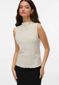 Haut sans manches crème en tissu texturé, avec un col en vagues et un ourlet brut. Associé à une jupe noire, le modèle pose devant un fond uni.