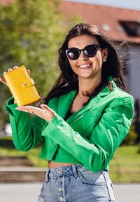 Yellow wallet with a snap closure in a smooth finish, held by a person wearing a green blazer and black sunglasses. Outdoor setting.