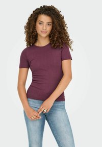 Kurzärmliges, tailliertes T-Shirt aus dunkelviolettem Rippstoff, kombiniert mit hellblauen Denim-Jeans. Bemerkenswerte Details sind die dezente Textur und das Muster.