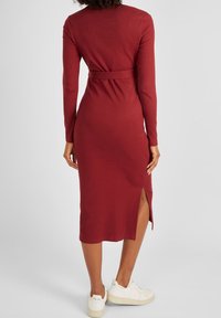 Langes, geripptes maroones Kleid mit Taillengürtel und seitlichem Schlitz, kombiniert mit weißen Sneakers. Das Kleid hat lange Ärmel und eine figurbetonte Silhouette.