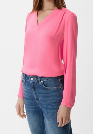 Kvinde iført en knaldpink bluse med lange ærmer og V-udskæring kombineret med blå denimjeans på en ensfarvet baggrund.