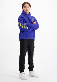 Paarse hoodie met gele bliksemschichtgraphics op de mouwen, gedragen met zwarte cargo-broeken en witte sneakers.