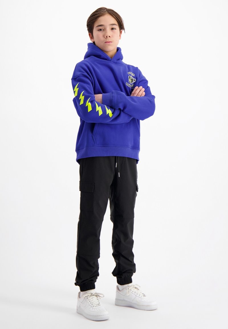 Paarse hoodie met gele bliksemschichtgraphics op de mouwen, gedragen met zwarte cargo-broeken en witte sneakers.