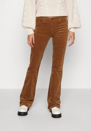 Personne portant un pantalon flare en velours côtelé marron, des bottes blanches épaisses et un pull en grosse maille crème avec des manches ballon.