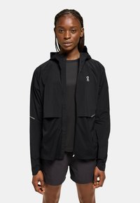Svart lättviktsjacka med huva, dragkedja framtill, elastiska manschetter och reflexdetaljer. Kombineras med en mörk topp och shorts, minimalistisk design.