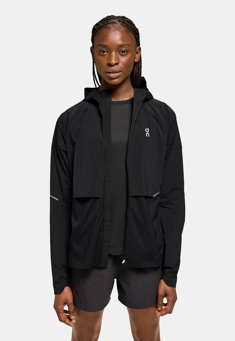 Svart lättviktsjacka med huva, dragkedja framtill, elastiska manschetter och reflexdetaljer. Kombineras med en mörk topp och shorts, minimalistisk design.