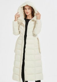 Manteau long et rembourré en beige clair avec une capuche. Présente un motif matelassé, une fermeture éclair à l'avant et des poches latérales, fabriqué en matériau synthétique lisse.