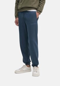 Pantalones de chándal azul marino con cintura elástica y cordón, que presentan un sutil logo de Calvin Klein. Textura suave y lisa; corte relajado.