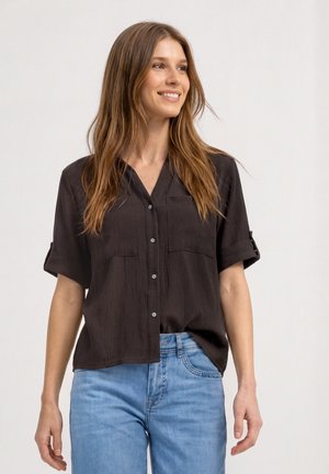 Femme portant une chemise noire à manches courtes boutonnée avec des poches et un jean bleu clair, souriante et regardant sur le côté sur un fond uni.