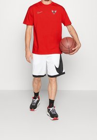 Hombre sosteniendo un balón de baloncesto que lleva una camiseta roja de los Chicago Bulls, pantalones cortos blancos de Nike, calcetines negros y zapatillas de baloncesto negras con suelas rojas.