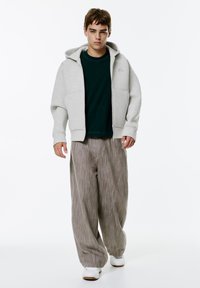 Sivá mikina na zips s kapucňou, nosená cez tmavozelené tričko. Voľné pruhované béžové nohavice a biele tenisky dopĺňajú outfit.