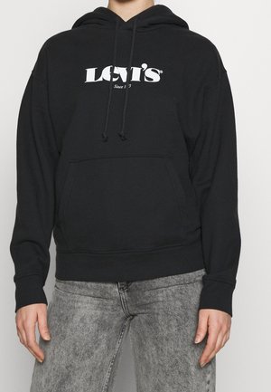 Personne portant un sweat à capuche noir Levi’s avec un logo blanc et un jean gris clair délavé, debout, les mains détendues le long du corps.