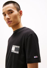 Camiseta de algodón negra con un bolsillo de parche con un diseño geométrico en blanco y negro. Cuello acanalado y detalle del logotipo en la manga.
