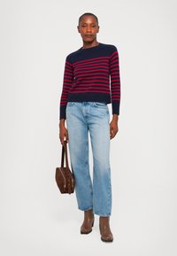 Tummansininen ja punaraitainen neuletakki, jossa on pyöreä pääntie, yhdistettynä vaaleansinisiin leveälahkeisiin farkkuihin ja ruskeisiin nilkkureihin. Ruskea käsilaukku kädessä.