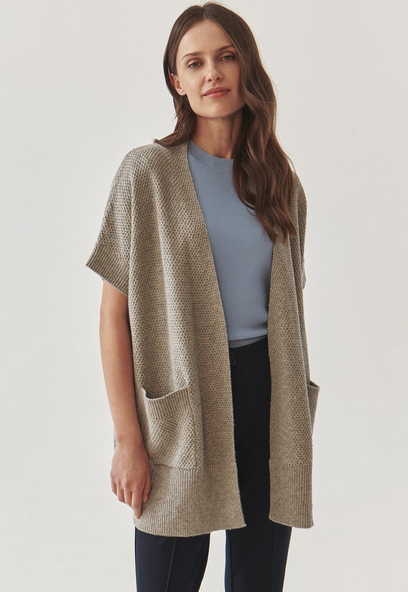 Cardigan en tricot beige clair avec des manches courtes, ouverture devant et deux poches avant, associé à un haut bleu et un pantalon foncé. Texture lisse.
