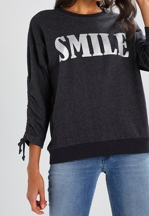 Camiseta de manga larga gris oscuro con "SMILE" en grandes letras blancas, mangas fruncidas con cintas, combinada con vaqueros azul claro.