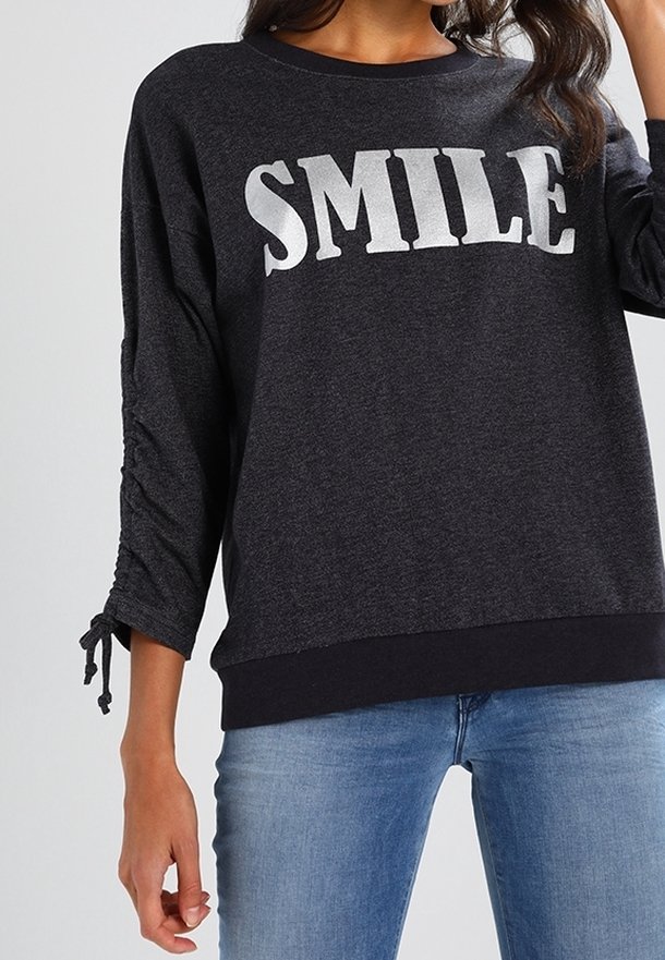 Chemise gris foncé à manches longues avec "SMILE" en grandes lettres blanches, manches froncées avec liens, portée avec un jean bleu clair.
