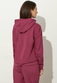 Felpa color bordeaux con un ampio cappuccio, polsini a costine e una vestibilità rilassata, realizzata in un morbido tessuto testurizzato. Vista dal retro su uno sfondo chiaro.