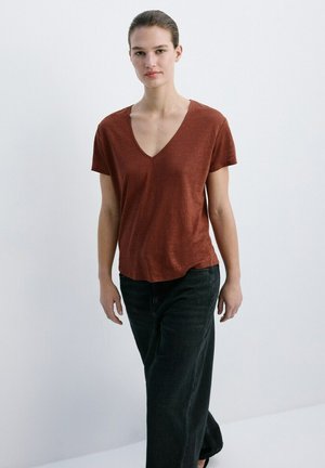 Tânără purtând un tricou scurt cu mâneci și decolteu în V, de culoare ruginie, și pantaloni largi negri, stând pe fundal alb simplu.