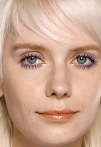 Close-up van een gezicht met lichte huid, kort platinumblond haar, blauwe ogen en paarse mascara op de wimpers, met glanzende nude lippen.