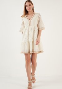 Robe courte en crème avec un décolleté en V, des boutons décoratifs et des manches volantées. Présente des broderies à œillets et un ourlet en étages avec des bords beiges.