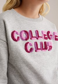 Sivka majica s kapuco z dvignjenimi črkami v večbarvnih odtenkih, ki črkujejo "KOLEGIJSKI KLUB" v različnih odtenkih roza in vijolične. Mehka tekstura.